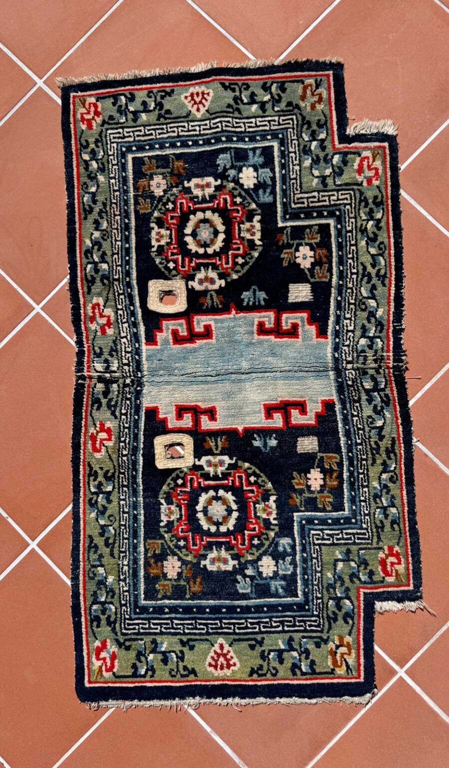 Un tappeto rettangolare a motivi ornamentali con due medaglioni floreali rossi e bianchi su sfondo scuro, bordo verde e disegni geometrici giace su un pavimento in cotto. Questo pezzo di Bozza automatica presenta una forma irregolare e un'usura visibile.