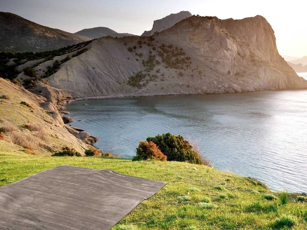 Un tappeto di bambù si trova su una collina erbosa che si affaccia su una baia tranquilla, con scogliere e montagne frastagliate sullo sfondo, sotto un cielo limpido.