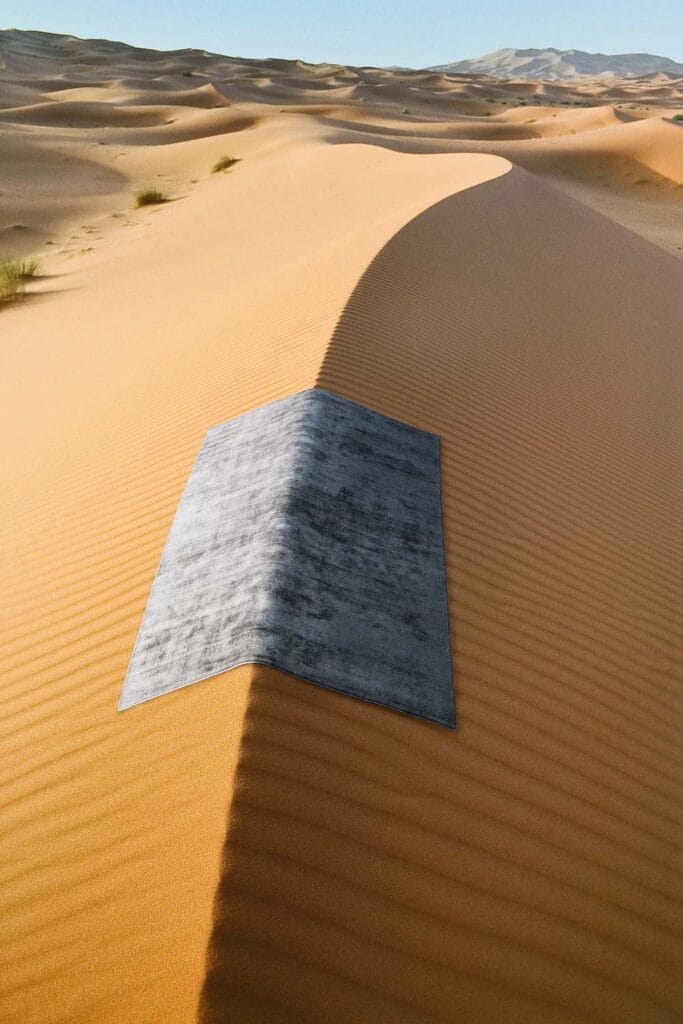 Un tappeto rettangolare grigio, come un'automatica Bozza, è posto ordinatamente sulla cresta di una duna di sabbia in un vasto paesaggio desertico, proiettando un'ombra netta lungo la curva della duna sotto un cielo limpido.