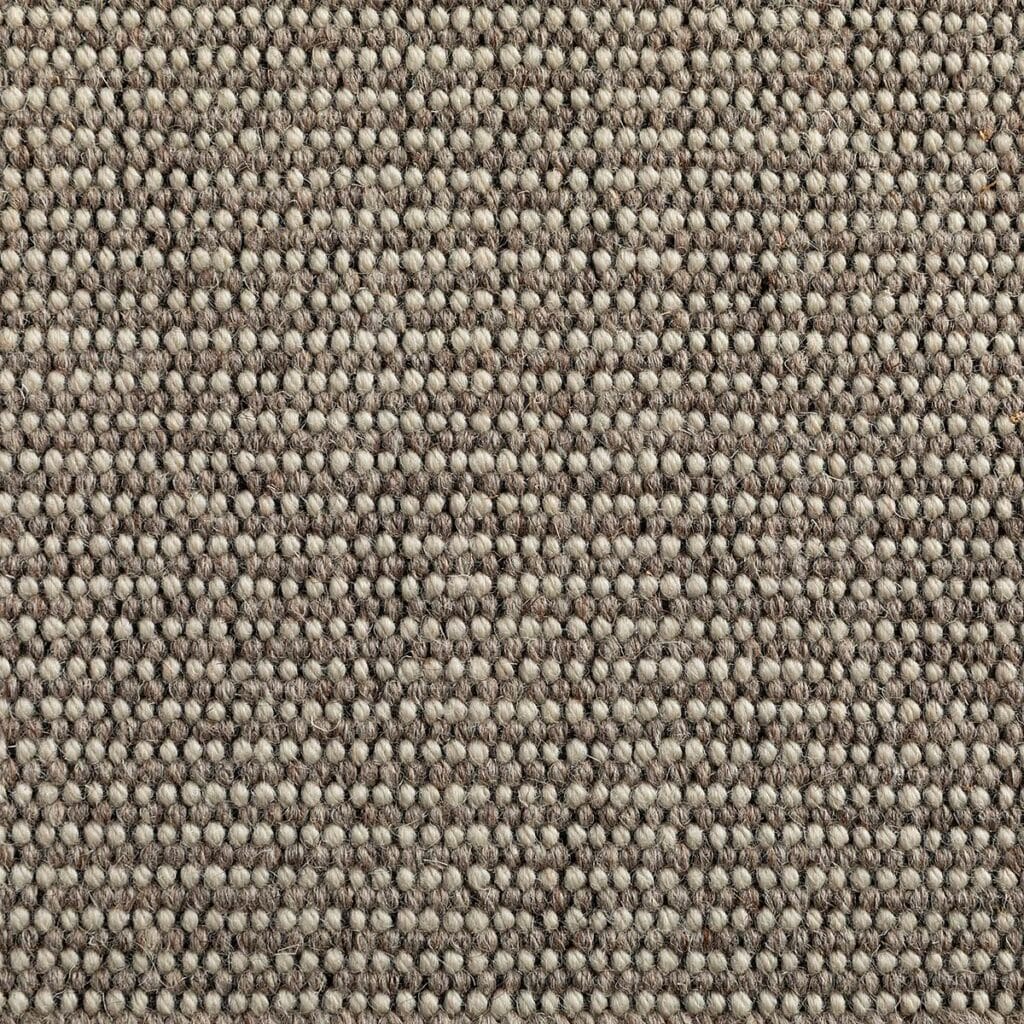 Primo piano di un tappeto testurizzato e intrecciato nei toni neutri del beige e del marrone, che mostra un motivo di fibre arrotondate e fitte, perfette per aggiungere un sottile calore a qualsiasi spazio. La tecnologia Bozza automatica ne migliora la durata e la finitura.