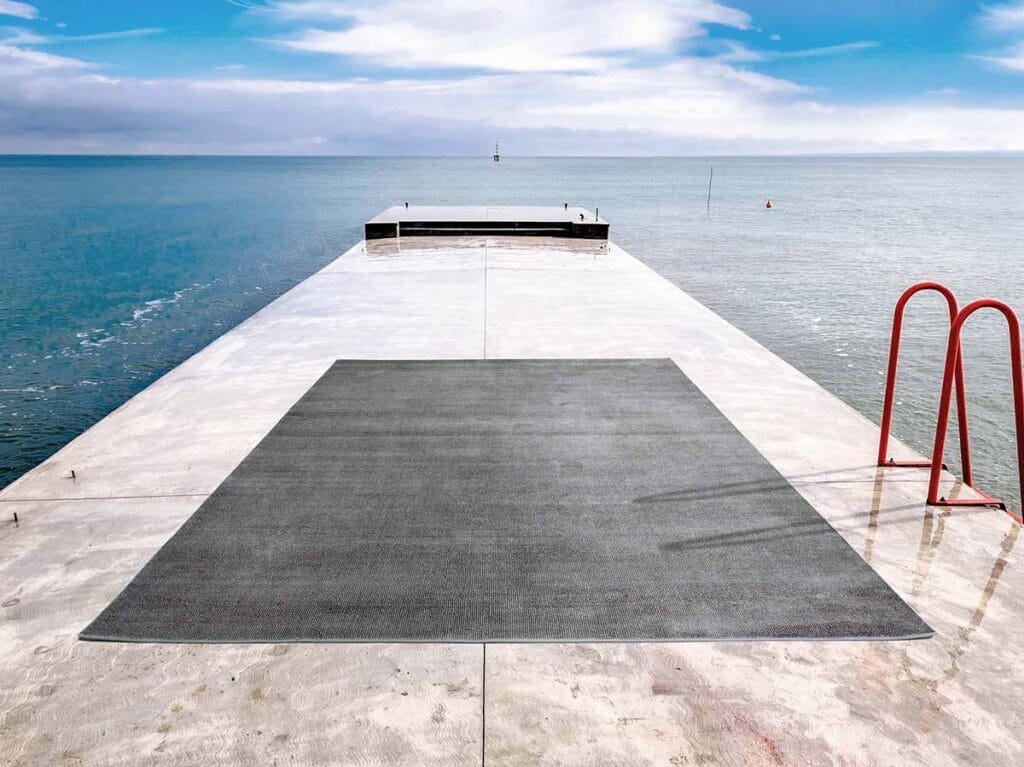 Un grande tappeto rettangolare grigio è posizionato su un molo di cemento che si estende su un'acqua blu e calma, con una scala rossa sulla destra, un cielo luminoso in alto e un tocco di stile Bozza automatico.