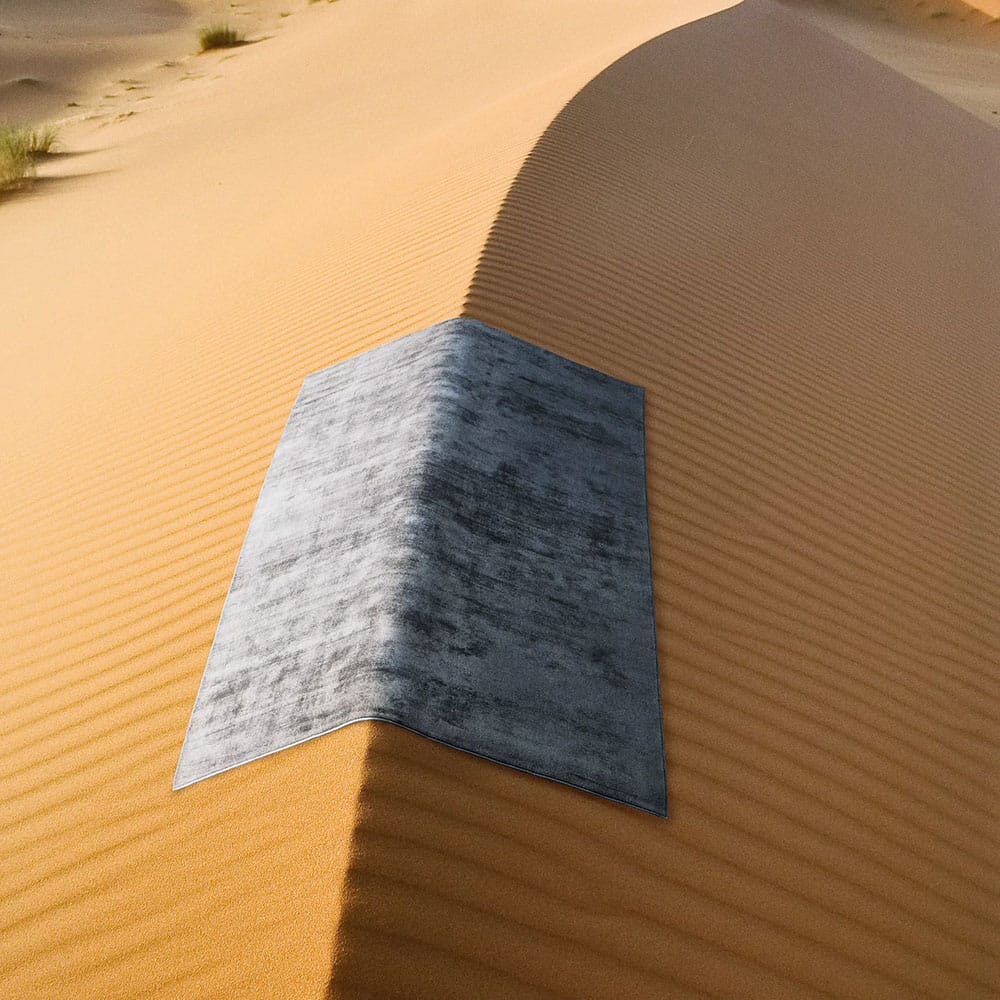Una moquette grigia e rettangolare è posata ordinatamente sulla cresta di una duna nel deserto, in netto contrasto con la sabbia liscia e arancione e con i disegni increspati delle dune.